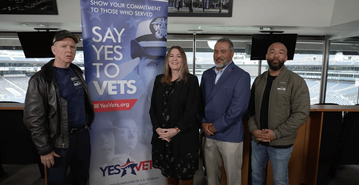 Four individuals stand next to a “Say Yes to Vets” banner at an indoor stadium suite, promoting veteran employment through the YesVets program. The group includes a mix of business and military-affiliated representatives, showing their commitment to hiring and supporting U.S. veterans.