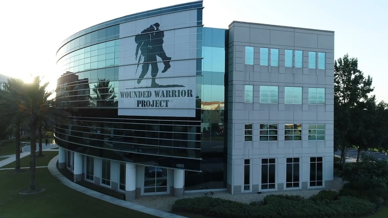 A modern office building with a large Wounded Warrior Project logo on its glass facade, surrounded by palm trees and a well-maintained landscape.