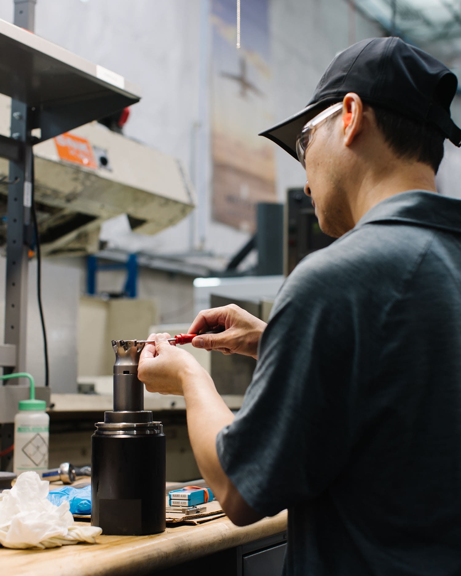 Employee in cap and safety glasses working on a machine part at a work bench.
