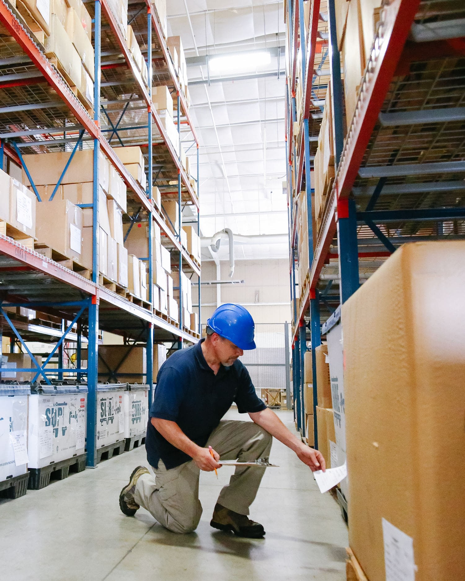 Man in a warehouse wearing a blue hard hat crouching and working with inventory on shelf.