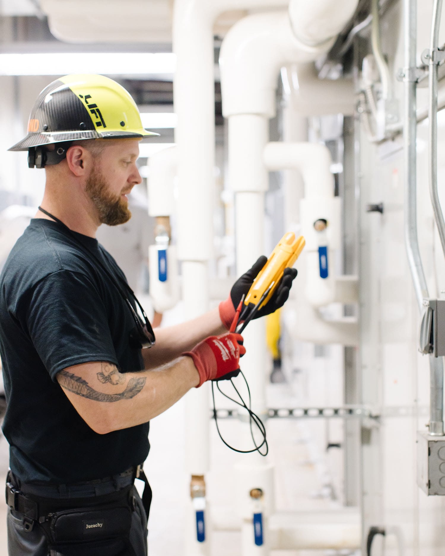 An employee in a yellow and black hard hat works with a multimeter device in a large utility room.