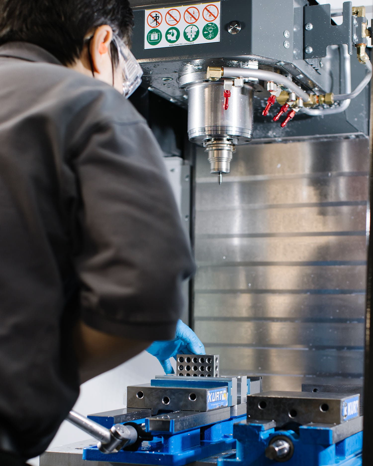 A worker in safety goggles and blue disposable gloves works with a sophisticated looking machine to fashion a rectangular metal component with holes drilled in it.