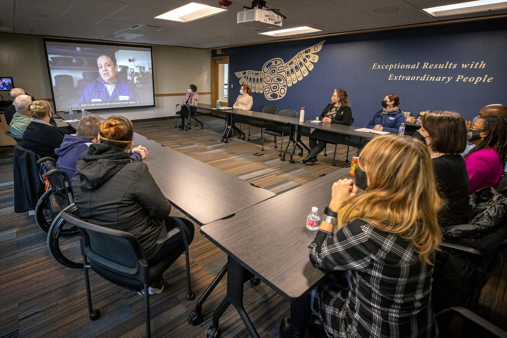 A group of attendees, including individuals in wheelchairs, watch a presentation in a conference room with a large screen displaying a speaker. The room features a mural with the words “Exceptional Results with Extraordinary People.”