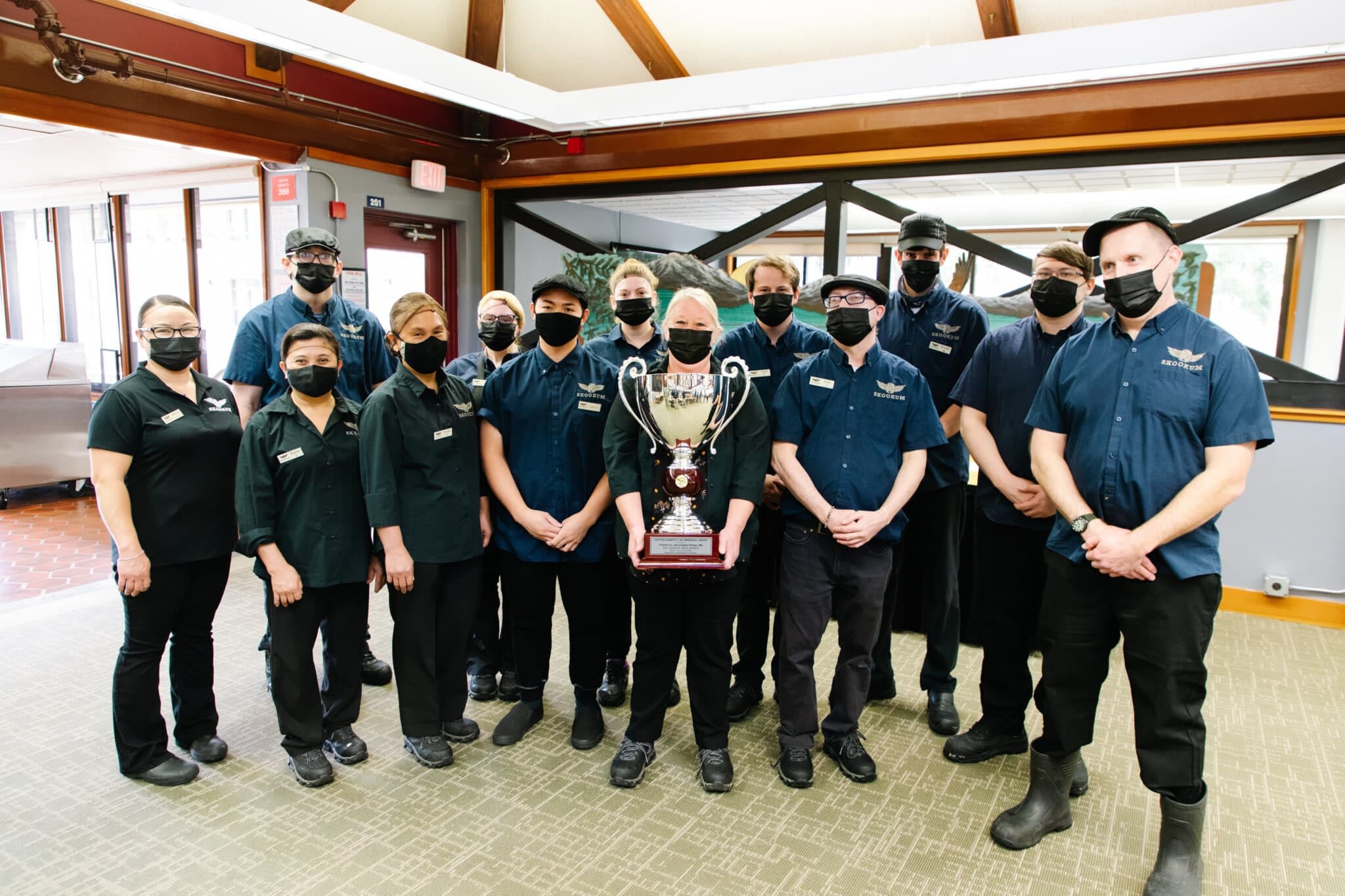 a group of eleven Skookum food service employees standing together inside a dining facility. They are all wearing navy blue uniforms with Skookum name tags and black face masks. The central figure, a woman, is holding a large silver trophy with ornate handles and a wooden base, signifying their achievement. The team members are standing close together, looking at the camera with a sense of pride. The background features wooden beams, large windows, and a mural of an eagle, creating a warm and welcoming atmosphere.