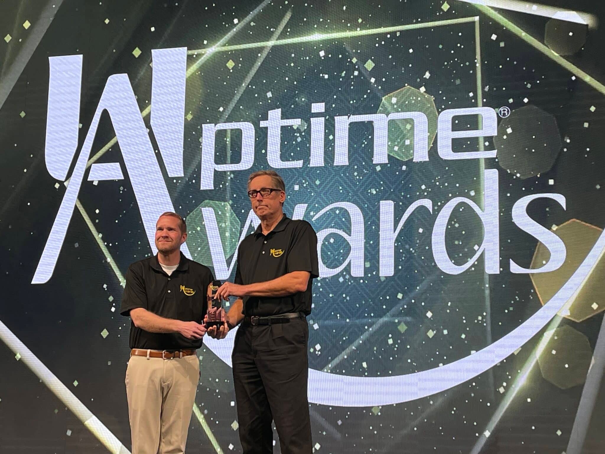 Two representatives from Tessera accept the 2023 Uptime Award for Best Digitalization for Reliability and Asset Management (RAM) on stage at the International Maintenance Conference. The backdrop features the Uptime Awards logo with a gold and black design.