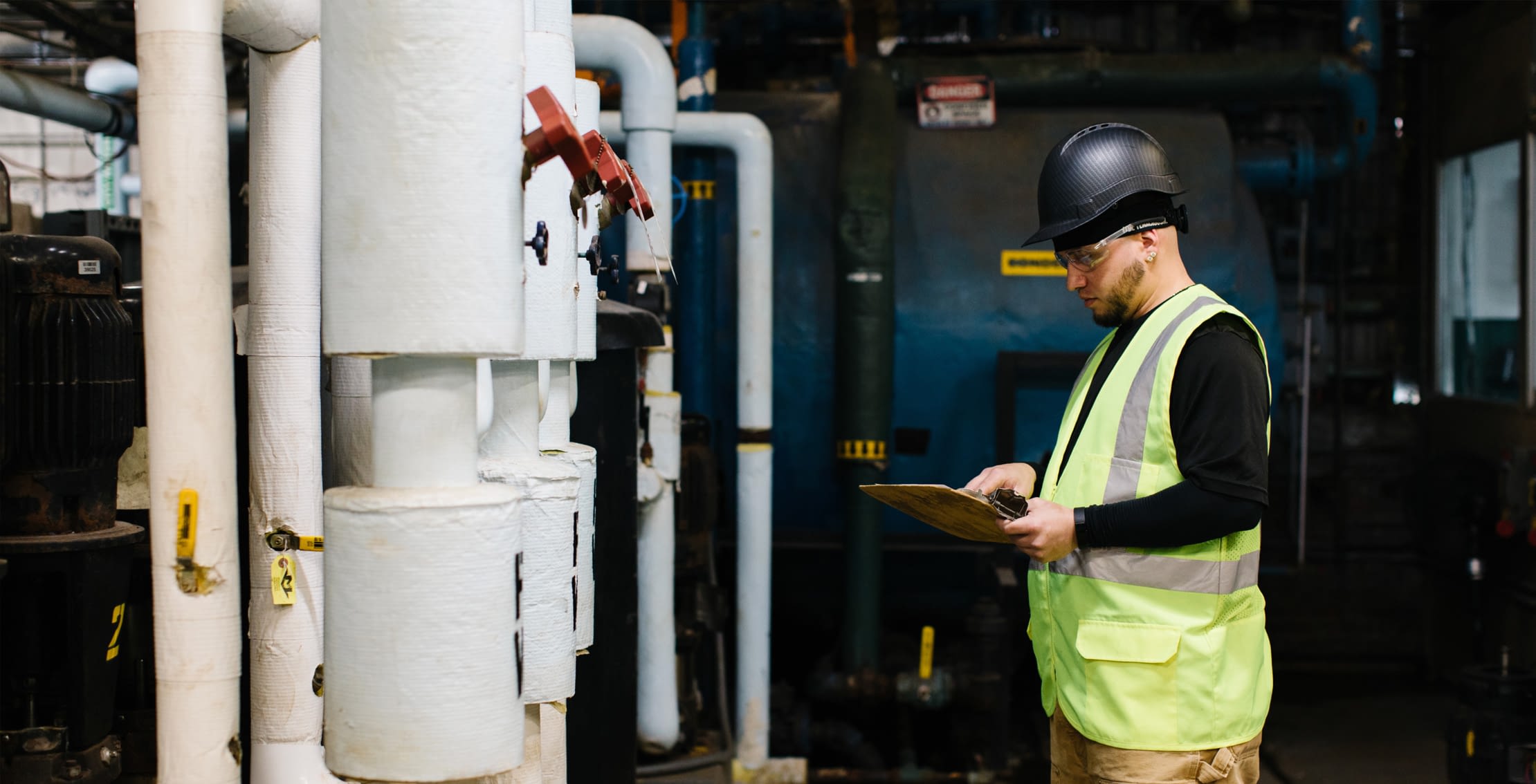 Inspector with hard hat and high visibility vest consult clipboard while inspecting industrial equipment.