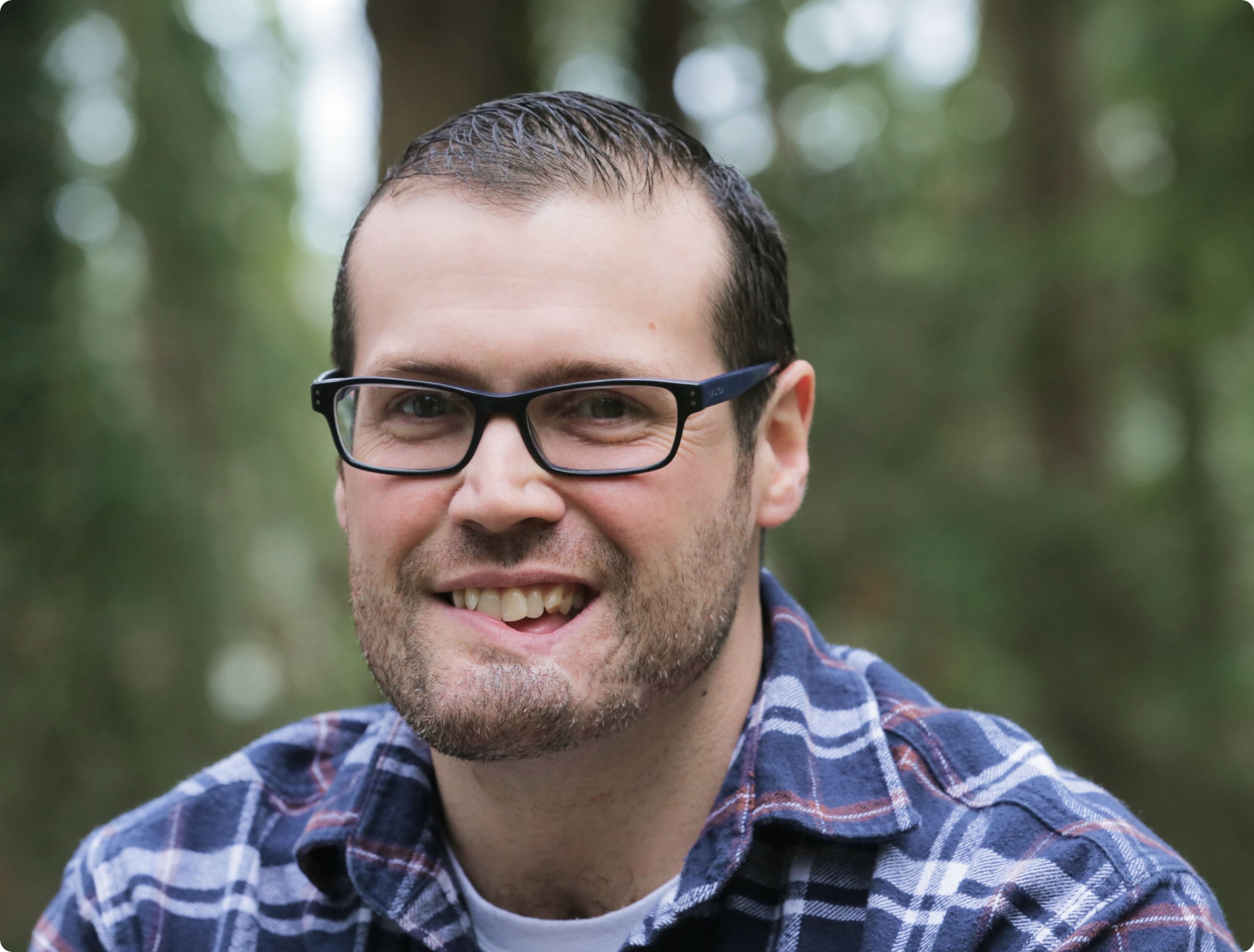 Man in glasses and plaid shirt smiling for the camera in front of the woods.