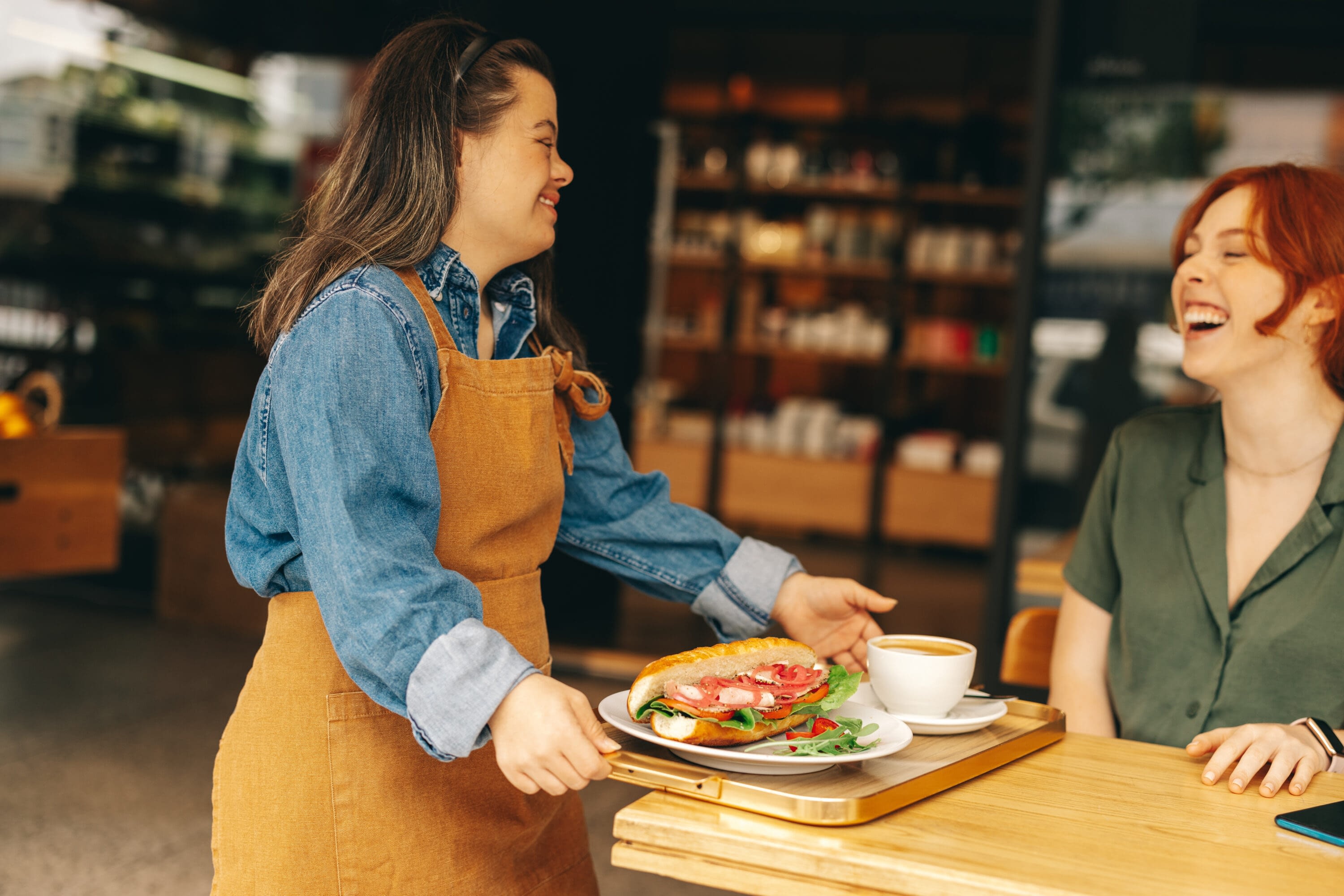 A friendly barista wearing a denim shirt and brown apron serves a happy customer at a local café. With a warm smile, she carries a tray with a fresh sandwich, salad, and coffee. The scene captures the joy of meaningful employment, highlighting the importance of inclusive workplaces that foster connection and opportunity within the community.