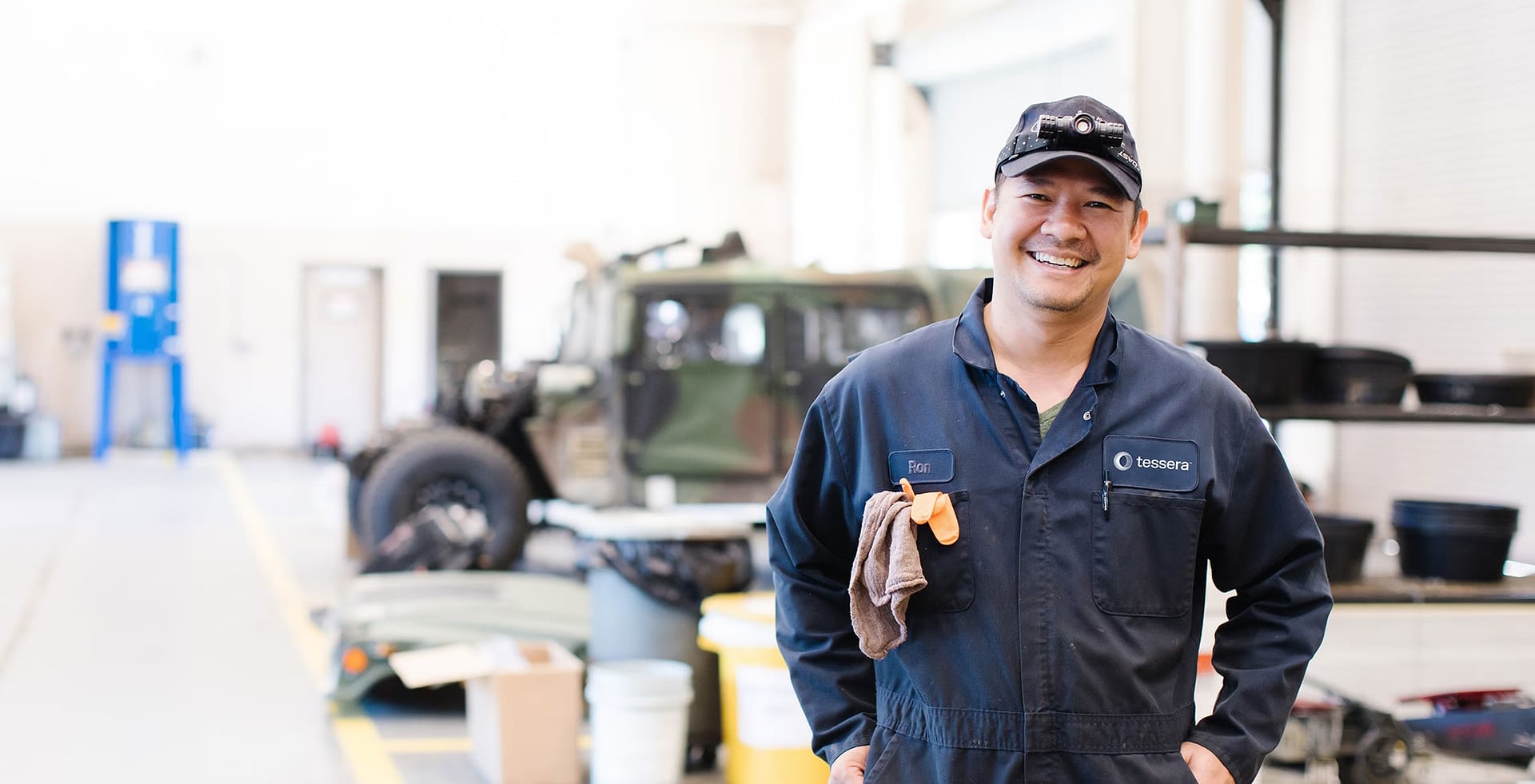 A Tessera mechanic in a navy-blue uniform and cap works under the hood of a vehicle, using tools to make adjustments. The setting is an industrial garage, with various equipment and tools visible in the background.