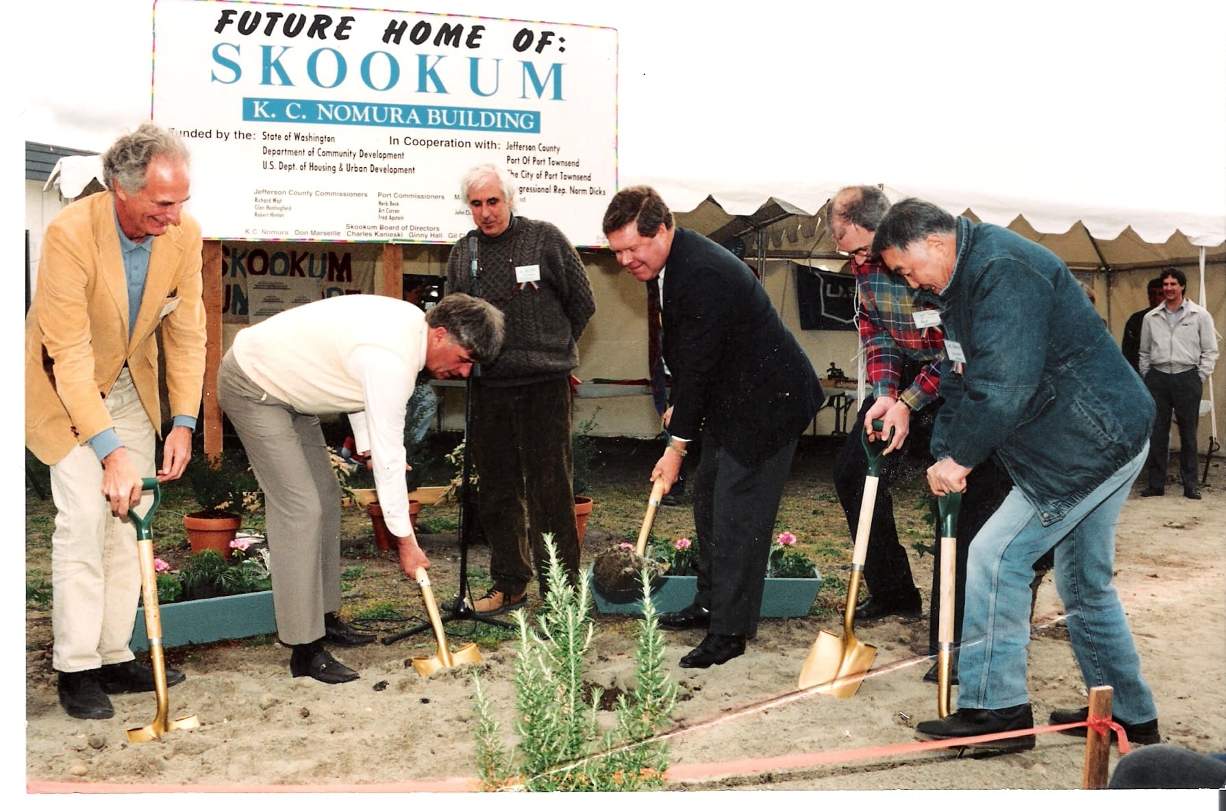A historical groundbreaking ceremony for the Skookum K.C. Nomura Building, featuring six men using golden shovels to turn the soil. A sign in the background reads "Future Home of: Skookum" and lists supporting organizations, including the State of Washington and Jefferson County. The event takes place under a tent, with onlookers observing in the background.