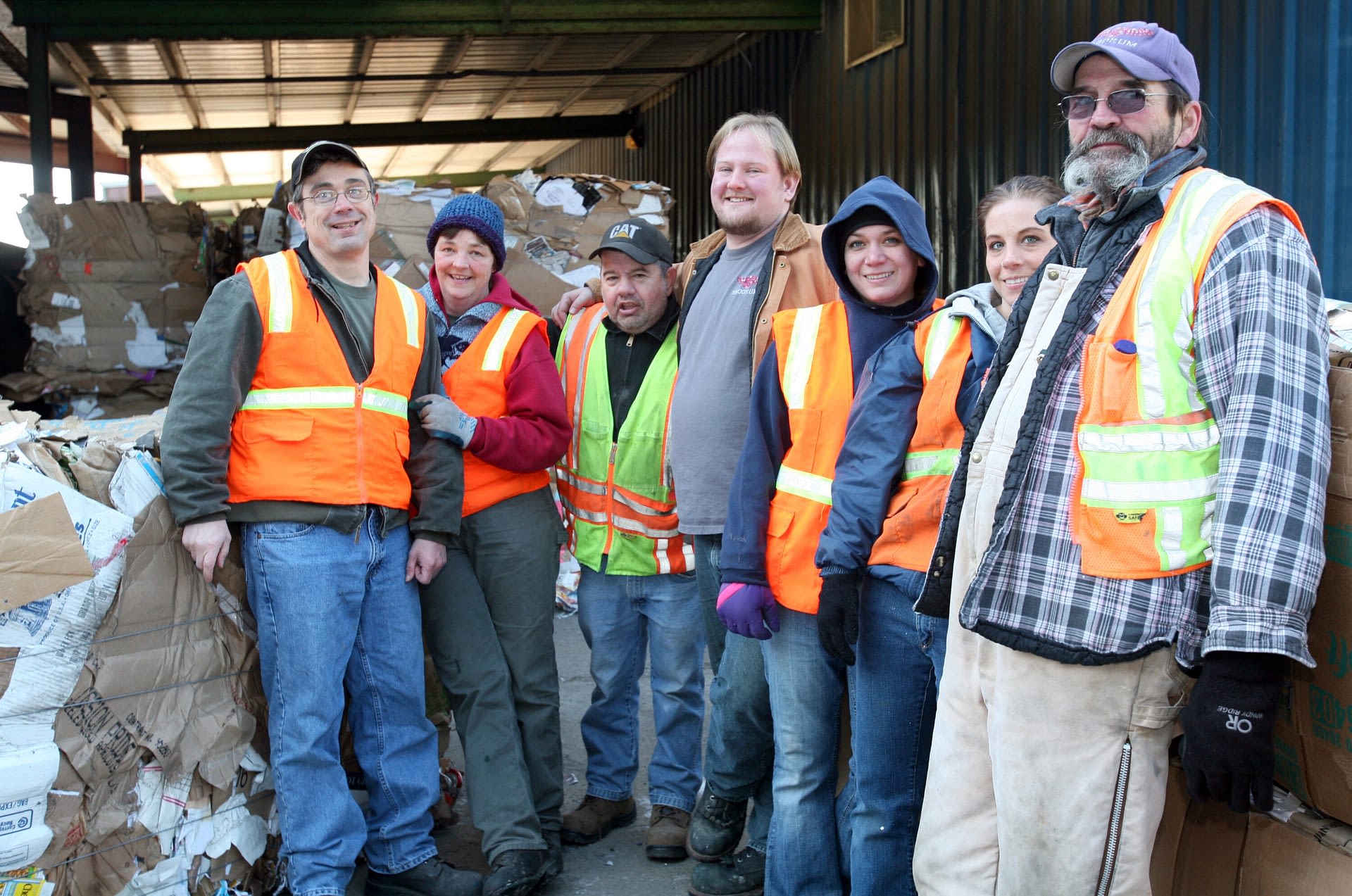 A group of seven smiling workers wearing reflective safety vests stand together in front of large stacks of compressed cardboard, likely at a recycling facility or warehouse. The individuals are dressed warmly, indicating a colder environment, and appear friendly and engaged.